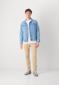 Lys blå denimjakke med knaplukning og to brystlommer, sammen med en hvid t-shirt, beige bukser og sko med grønne detaljer.