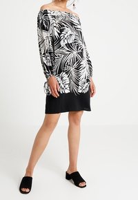 Nainen, jolla on yllään musta-valkoinen olkapäät paljastava mekko, jossa on lehtikuvio, pitkät hihat ja mustat avokkaat, joissa on paksu korko.