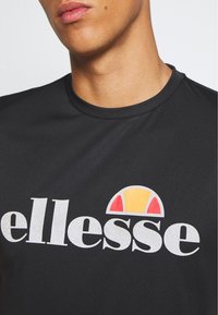 Černé tričko s krátkým rukávem s bílým logem "ellesse" a barevným půlkruhovým grafickým prvkem v žluté a červené nad textem.