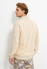 Beżowy sweter z wysokim kołnierzem, prążkowanymi mankietami i dolnym obszyciem. Model łączy go z beżowo-niebieskimi spodniami w kamuflaż.