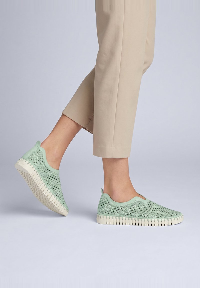 Persona che indossa pantaloni beige e sneaker slip-on verde chiaro con ritagli decorativi e suole bianche flessibili che cammina su uno sfondo semplice.