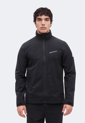 Junger Mann, der nach vorne schaut und eine schwarze Reißverschlussjacke mit Peak Performance-Logo auf Brust und Ärmel trägt, vor einem einfarbigen Hintergrund.