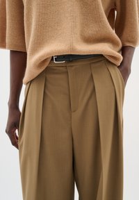Beige gerippter Strickpullover kombiniert mit braunen, hoch taillierten, plissierten Hosen. Mit einem schwarzen Gürtel mit einer silbernen Schnalle. Glatte Stoffoberfläche.
