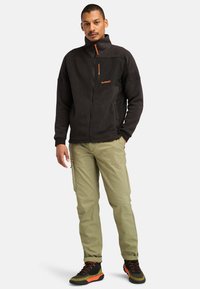 Giacca in pile nera con colletto alto, zip arancione a contrasto e logo; abbinata a pantaloni cargo verde chiaro e scarpe da trekking nere.