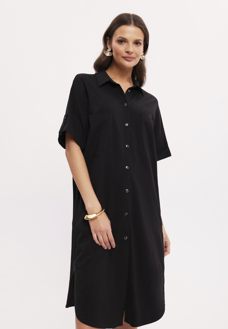 Czarna sukienka koszulowa z klasycznym kołnierzykiem, krótkimi rękawami, guzikami z przodu oraz dwiema kieszeniami na piersiach. Gładka tekstura i długość do kolan.