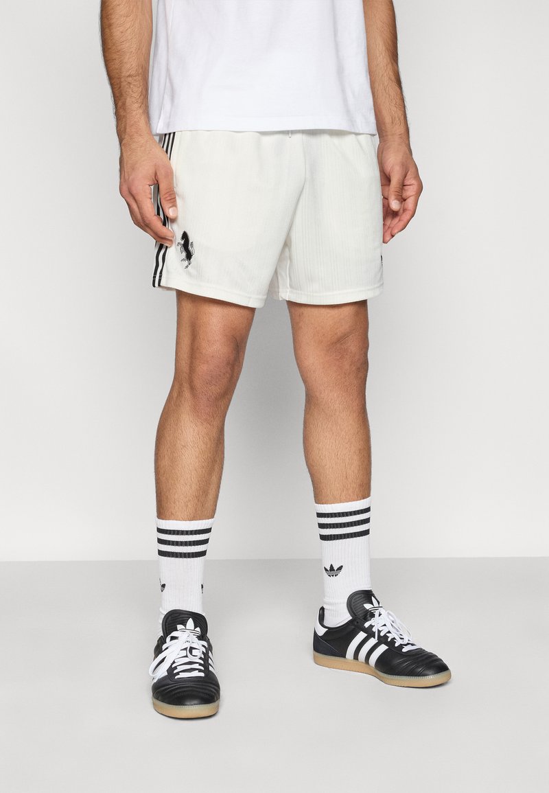 Weiße Sportshorts mit dezenten vertikalen Streifen, schwarzen Seitenakzenten und Logo. Kombiniert mit schwarzen Turnschuhen und weißen gestreiften Socken.