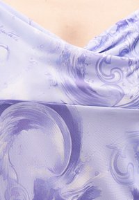 Κοντινό πλάνο ενός λιλά φορέματος με σπειροειδές σχέδιο και διακριτικό V-λαιμόκοψη, που δείχνει το πάνω μέρος του στήθους και τις λεπτομέρειες του υφάσματος.