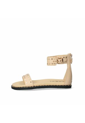 Infradito beige con suola piatta, cinturino alla caviglia, dettagli con borchie e suola nera texturizzata. Design semplice con accenti minimalisti.