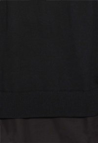 Schwarzer Strickpullover mit geripptem Saum und glatter Textur, kombiniert mit einem einfarbigen schwarzen Stoff darunter für ein schichtartiges Aussehen.