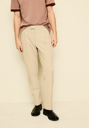 Mann trägt beige maßgeschneiderte Hose, schwarze Leder-Stiefeletten und ein kurzärmeliges rotes Polohemd mit Muster vor einem schlichten Hintergrund.