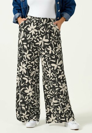 Czarne spodnie o szerokich nogawkach z wyrazistym, białym, abstrakcyjnym kwiatowym wzorem, noszone z białymi tenisówkami i niebieską jeansową kurtką. Ręce w kieszeniach.