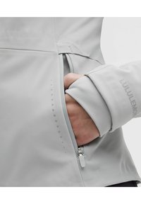 Chaqueta deportiva gris claro con bolsillo con cremallera, textura suave y logo en el puño. Patrón de puntos sutil a lo largo de la costura.