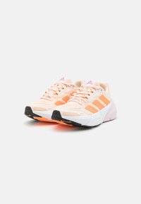 Baskets pêche clair avec trois bandes orange, dessus en maille texturée, languette rembourrée, et semelle extérieure en caoutchouc noir avec détail logo jaune.