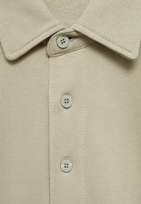 Beżowa koszulka polo z teksturowanym kołnierzykiem i trzema guzikami, wykonana z gładkiego dzianinowego materiału z delikatnymi detalami ściegów.