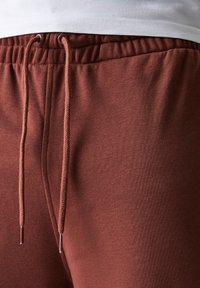 Pantaloni della tuta color ruggine con vita elastica, coulisse regolabile e texture liscia. Presentano una tasca laterale per maggiore comodità.