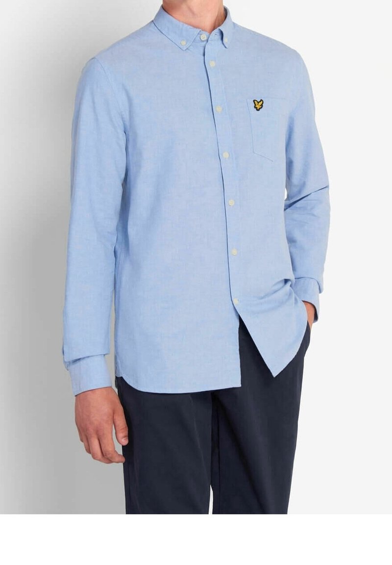 Hombre que lleva una camisa de manga larga de botones azul claro con un bolsillo en el pecho y pantalones oscuros, de pie con una mano en el bolsillo.