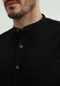 Chemise noire à col mao, ornée de boutons bleus et d'une texture lisse et mate. Vue en gros plan mettant en avant les détails du col.