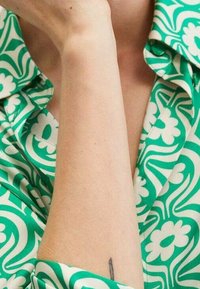 Blouse verte à motifs présentant de grandes fleurs et des dessins tourbillonnants. Le col est replié, avec un gros plan sur un bras nu et un tatouage discret.
