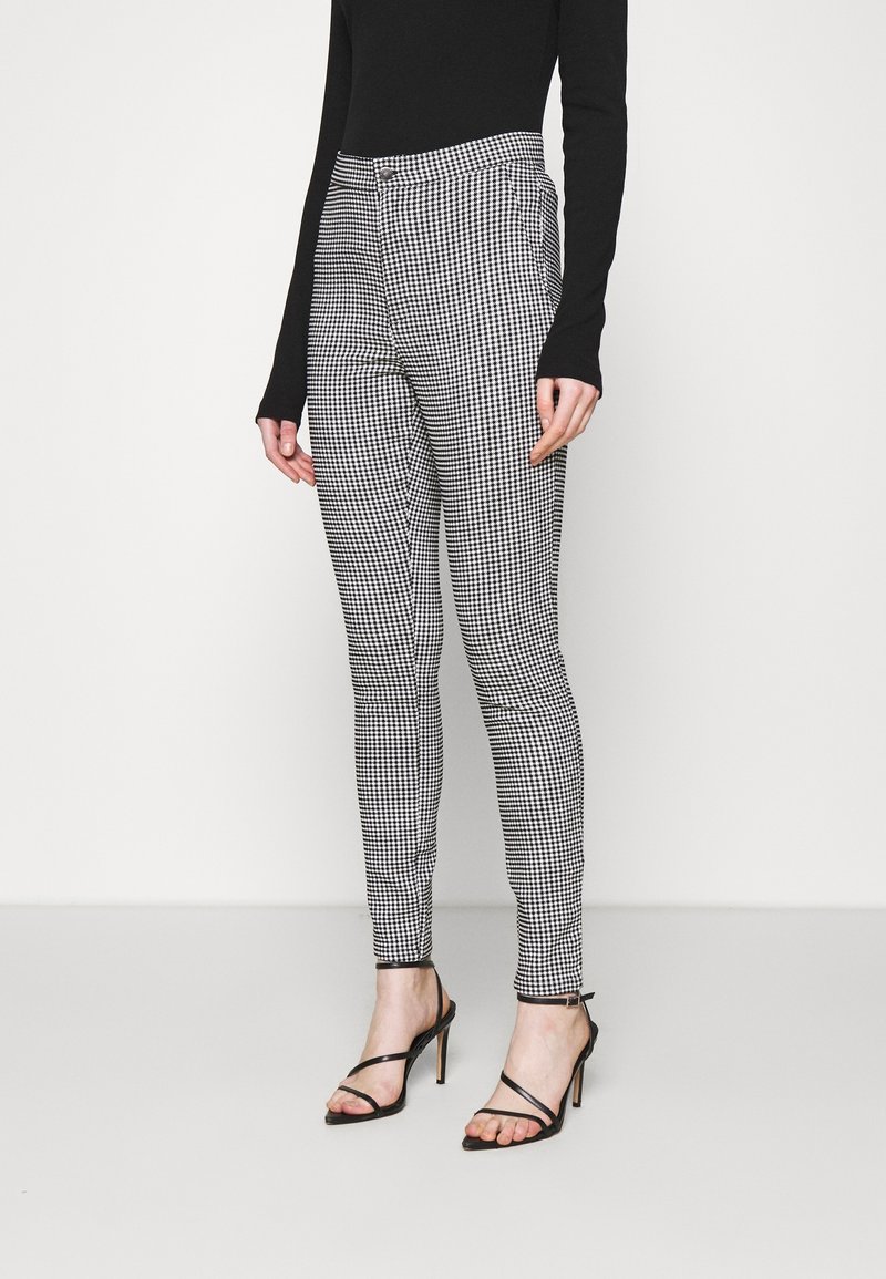 Femme portant un pantalon slim à carreaux noir et blanc, un haut noir à manches longues et des sandales à talons hauts noires à brides, debout sur un fond blanc.