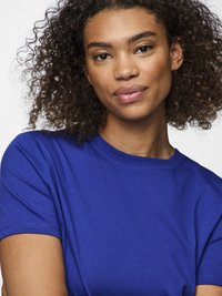 Mladić s kovrčavom kosom u jarko plavoj kratki rukavima, samouvjereno gleda u kameru na jednobojnoj bijeloj pozadini.