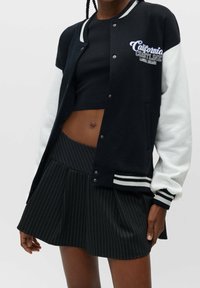 Black and white varsity jacket with ribbed cuffs and neckline, featuring "California Coastline" embroidery, paired with a pleated black pinstripe skirt.