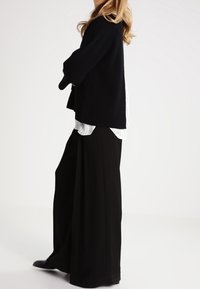 Schwarze gerippte Strickjacke über einem weißen gestreiften Hemd, kombiniert mit weit geschnittenen schwarzen Hosen. Das Outfit zeichnet sich durch einen lässigen Schnitt und weiche Texturen aus.