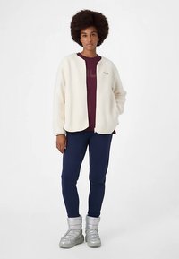 Mjuk krämfärgad fleecetröja över en vinröd skjorta, kombinerad med marinblå byxor och metallicsölva stövlar, med en minimalistisk logga på jackan.