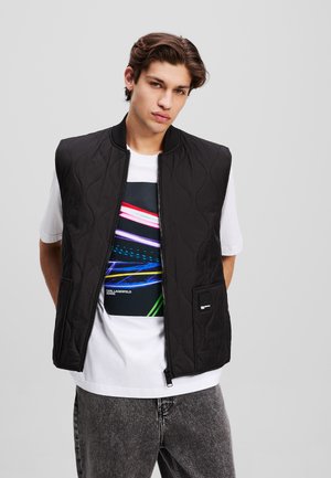 Gilet nero trapuntato con collo a costine, chiusura con zip e due tasche laterali. Indossato sopra una maglietta bianca con grafica che presenta linee astratte colorate.