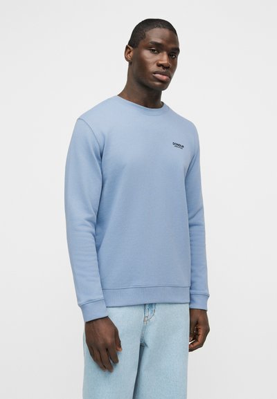 Άνδρας που φοράει γαλάζιο φούτερ Dondup και γαλάζια τζιν, στέκεται μπροστά σε απλό λευκό φόντο.