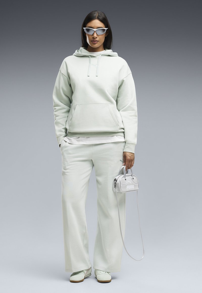 Γυναίκα με ανοιχτό πράσινο hoodie και παντελόνι με φαρδιά μπατζάκια, φοράει γυαλιά ηλίου, κρατάει μικρή τσάντα Puma και στέκεται μπροστά σε απλό φόντο.