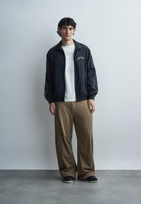 Junger Mann mit lockigem dunklem Haar, der eine schwarze Jacke mit Reißverschluss, ein weißes T-Shirt, weite braune Hosen und schwarze Turnschuhe vor einer schlichten grauen Wand trägt.