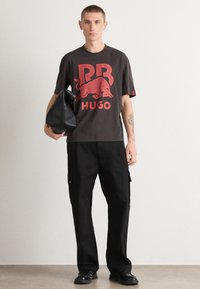 Black T-shirt with bold red "RB HUGO" logo, paired with black cargo pants and a black handbag. Smooth textures and relaxed fit.