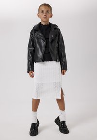 Giovane ragazza che indossa una giacca di pelle nera, top nero, gonna bianca lavorata a maglia con spacco laterale, calzini bianchi e mocassini neri, in piedi davanti a uno sfondo semplice.