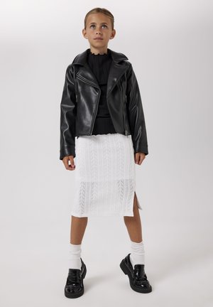 Giovane ragazza che indossa una giacca di pelle nera, top nero, gonna bianca lavorata a maglia con spacco laterale, calzini bianchi e mocassini neri, in piedi davanti a uno sfondo semplice.