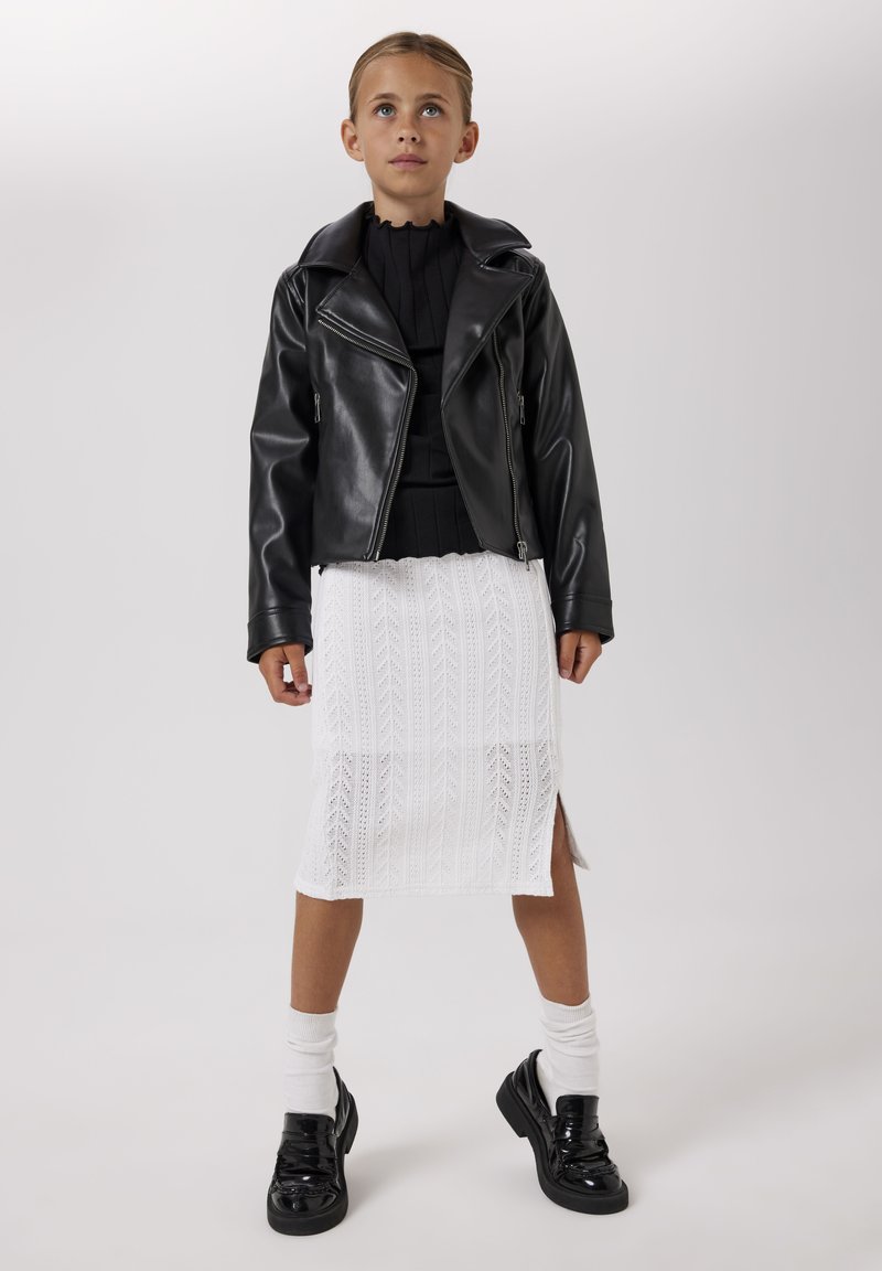 Giovane ragazza che indossa una giacca di pelle nera, top nero, gonna bianca lavorata a maglia con spacco laterale, calzini bianchi e mocassini neri, in piedi davanti a uno sfondo semplice.