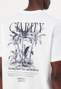 Balta medvilnės marškinėliai su dideliu juodu "CLARITY" spaudiniu, esančiu virš išsamaus mėlyno botaninio piešinio, ir tekstu nugarėlėje.