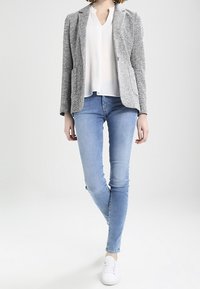 Veste grise texturée portée sur un chemisier blanc, associée à un jean skinny bleu clair et des baskets blanches. Présente une coupe ajustée et des lignes épurées.