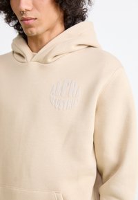 Felpa beige con tasca a marsupio, caratterizzata da un logo circolare testurizzato che recita "ALPHA INDUSTRIES" sulla parte frontale. Materiale in morbido cotone.