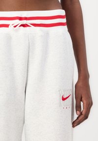 Grå joggingbyxor med röd och vit randig midja, med en röd Nike-logotyp och textilstruktur. Inkluderar ett dragsnöre för justerbarhet.
