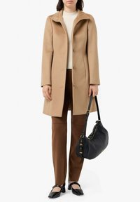 Cappotto beige in misto lana con colletto alto e bottoni frontali, abbinato a pantaloni marroni e una borsa a spalla in pelle nera. Le ballerine nere completano il look.