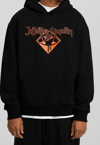 Felpa con cappuccio nera con grafica arancione che mostra il testo "JuJutsu Kaisen" e una forma a diamante. Il materiale sembra morbido, con una tasca frontale.