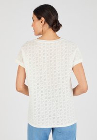 Witte kortemouwtop met een textuurpatroon van kleine gaatjes en blauwe en rode accenten, met een ronde halslijn en een relaxed fit.