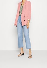Blazer rosa sopra una blusa a fantasia, abbinato a jeans cropped blu chiaro e sandali neri con tacco. Il tessuto appare liscio e morbido.