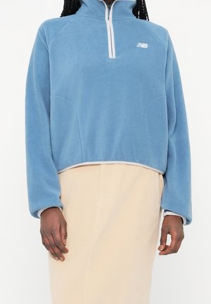 Person trägt einen hellblauen Fleece-Pullover mit weißem Reißverschluss und Logo, kombiniert mit einem beigen Rock vor weißem Hintergrund.