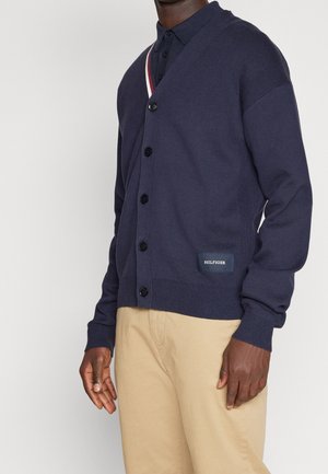 Marine gebreide cardigan met een v-hals, knoopsluiting, geribbelde boorden en tailleband. Voorzien van een klein logopatch aan de onderzijde van de voorkant.