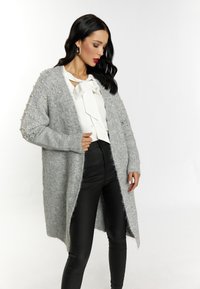 Cardigan lungo grigio realizzato in un morbido materiale peloso, arricchito da dettagli in perle sulle maniche, indossato sopra una camicia bianca e pantaloni neri.