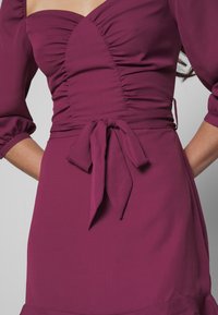 Bordeauxklänning med en figurnära byst, puffärmar och ett knutet midjeband. Slät, mjuk tygkvalitet med subtil rynkning över toppen.