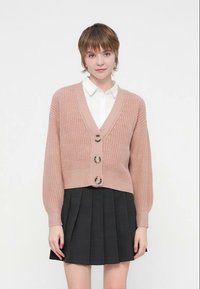 Różowy, dziany kardigan z dekoltem w serek, zapinany na guziki z prążkowanym dołem i bufkami na rękawach, zestawiony z plisowaną czarną spódnicą.