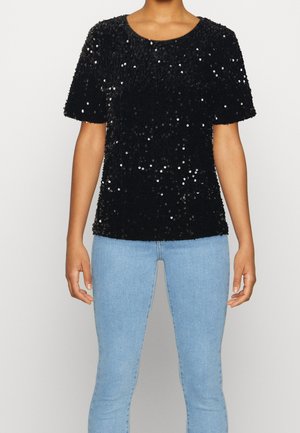 Kvinde iført en sort kortærmet paillet top og lyseblå jeans, stående mod en ensfarvet baggrund.