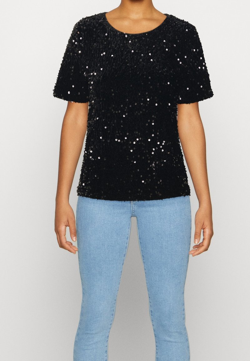 Femme portant un haut noir à manches courtes avec des sequins et un jean bleu clair, debout devant un fond uni.